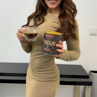 Mulher com vestido bege segura chávena de vidro e recipiente de suplemento Focus junto a móvel preto e parede branca com espelhos.