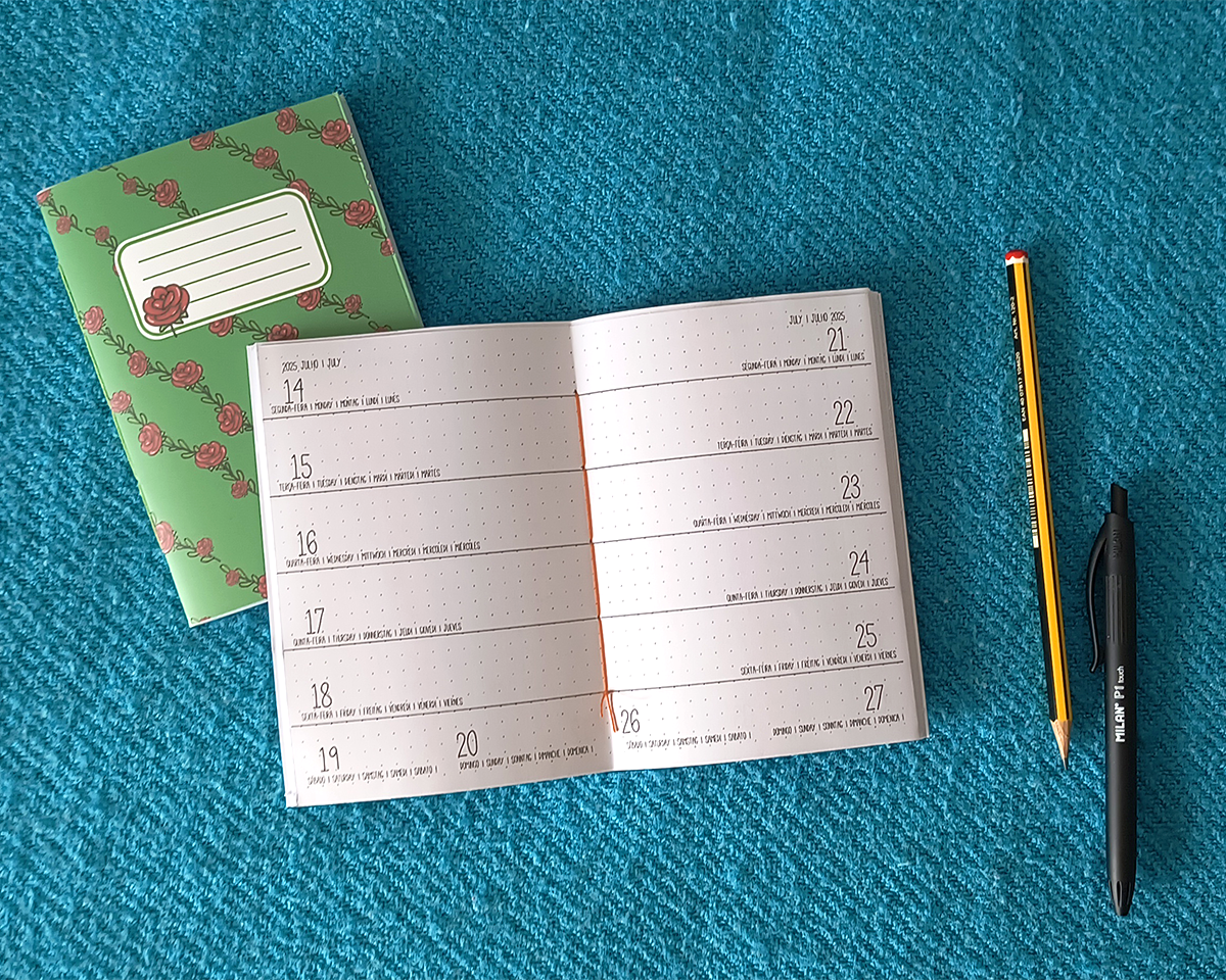 Agenda de Bolso A6 (Semanal) caderno verde com flores, calendário semanal aberto, lápis e caneta sobre tecido azul
