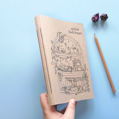 Caderno de capa kraft com ilustração preta e texto, lápis e dois dados em fundo azul claro