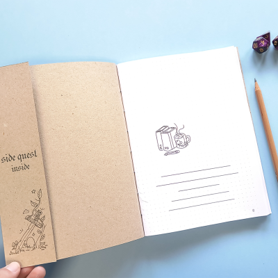 caderno aberto com capa marrom, lápis e dois dados roxos sobre superfície azul