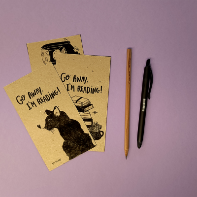 Dois marcadores de livros em papel castanho com ilustrações e texto, lápis e caneta numa superfície roxa