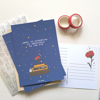 Conjunto de papelaria com cartões azuis e cartão postal com flor, fita adesiva vermelha e canetas em fundo claro.