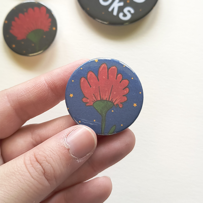 Botão redondo com flor vermelha e estrelas num fundo azul, segurado por mão, com outros botões ao fundo
