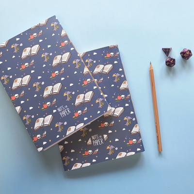 Dois cadernos azuis com padrão de livros, flores e texto, lápis e três dados coloridos sobre fundo azul claro.