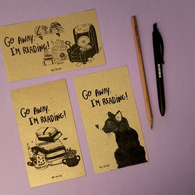 Três postais castanho claro com texto e desenhos a preto, ao lado lápis e caneta sobre fundo lilás