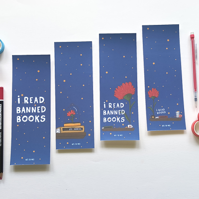 Marcadores de livros azuis com texto 'I READ BANNED BOOKS' e ilustrações coloridas, ao lado de lápis e fitas adesivas