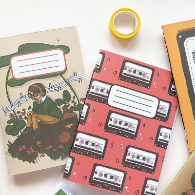 Três cadernos coloridos com capas decoradas: cassetes, ilustração de pessoa com auscultadores, texto 'MUSIC IS TO THE SOUL', caneta e fita adesiva.