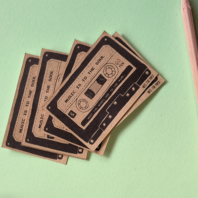 Cinco etiquetas castanhas com impressão de cassete e texto, sobre fundo verde com lápis e caneta ao lado.