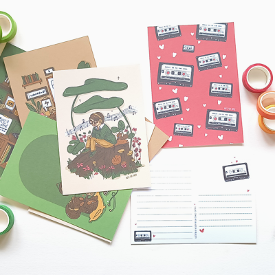 Cartões postais ilustrados com temas de natureza e cassetes, fitas adesivas coloridas e caneta sobre mesa branca.