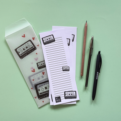 Lista de papel com desenhos de cassetes, envelopes transparentes com corações e três canetas sobre superfície verde