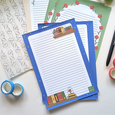 Papel de carta decorativo com linhas e ilustrações, fita adesiva colorida e canetas numa mesa branca.