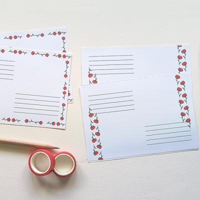 Postais com padrão de rosas vermelhas com caules verdes, caneta preta, lápis e fitas adesivas vermelhas numa mesa clara