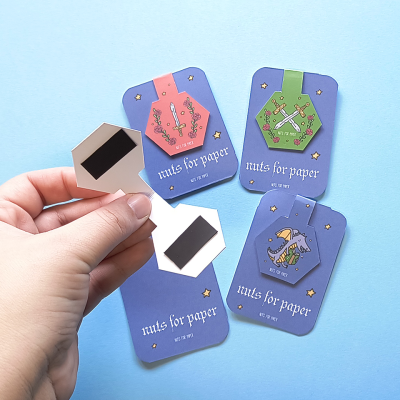 Marcadores magnéticos para papel com desenhos coloridos em cartão roxo e mão segurando dois marcadores hexagonais bege