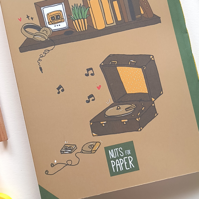 Caderno com capa ilustrada mostra gira-discos, disco, leitor, estante e o texto 'NUTS FOR PAPER'