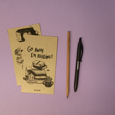 Dois cadernos castanhos com texto e desenhos, um lápis e uma esferográfica sobre fundo lilás