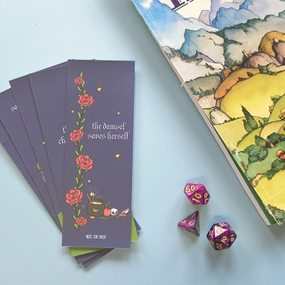 Marcadores de livro roxos com ilustrações e texto, três dados poliedros roxos com números dourados, e parte da capa de um livro ou jogo colorido.