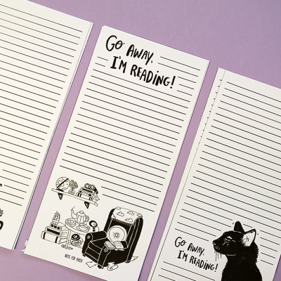 Três blocos de notas com desenhos de gatos, livros e texto 'Go Away! I'M READING!', sobre fundo lilás com lápis e caneta.
