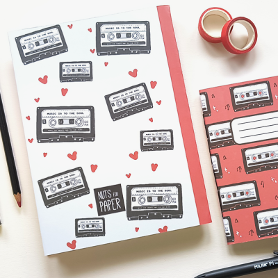 Dois cadernos com capas com desenhos de cassetes musicais e corações, acompanhado de material de escrita e fitas adesivas.
