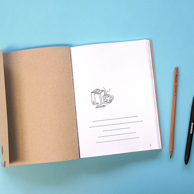 Caderno aberto com desenho de pacote de leite e chávena, lápis e caneta sobre fundo azul.