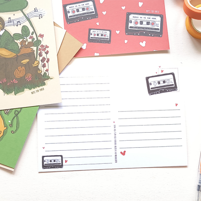 Papelaria com folhas de carta, cartões ilustrados, envelopes, fita adesiva e caneta laranja.