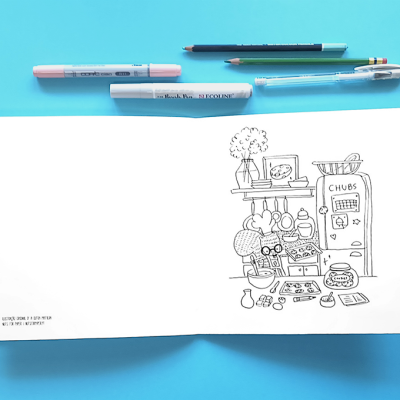 Livro de desenho com ilustração de cozinha em preto e branco e material de desenho azul e rosa