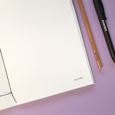 Caderno aberto com páginas pontilhadas, lápis e caneta sobre fundo lilás.