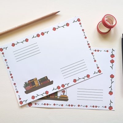Dois postais decorados com flores e ilustrações de livros sobre mesa branca com lápis, caneta e fita adesiva