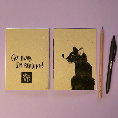 Dois cadernos com capa castanha reciclada, um com texto e outro com desenho de gato, ao lado de lápis e caneta sobre fundo rosa.