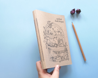 Caderno de capa kraft com ilustração preta e texto, lápis e dois dados em fundo azul claro