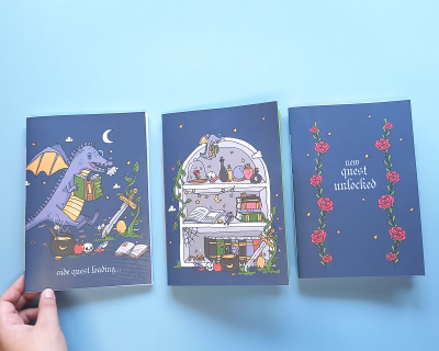 Três cadernos com capas azuis e desenhos de fantasia, incluindo dragão, poções e rosas.