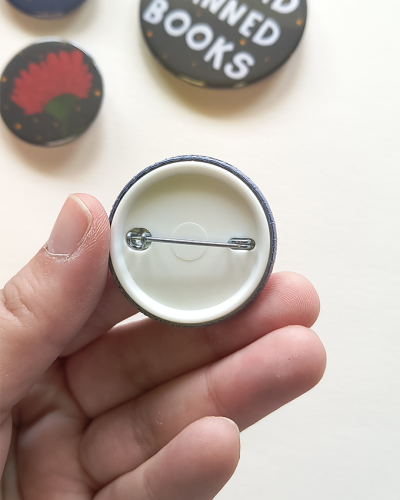 Botão redondo com alfinete visto por trás, segurado pela mão, com outros botões desfocados ao fundo