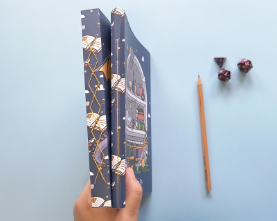 Livro de capa dura azul com ilustrações coloridas, lápis e dados políedricos