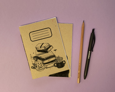 Cadernetas castanhas com ilustrações, lápis de madeira e caneta preta sobre superfície rosa