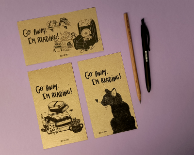 Três postais castanho claro com texto e desenhos a preto, ao lado lápis e caneta sobre fundo lilás