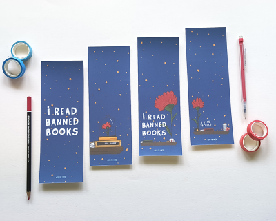 Marcadores de livros azuis com texto 'I READ BANNED BOOKS' e ilustrações coloridas, ao lado de lápis e fitas adesivas