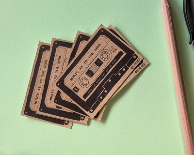 Cinco etiquetas castanhas com impressão de cassete e texto, sobre fundo verde com lápis e caneta ao lado.