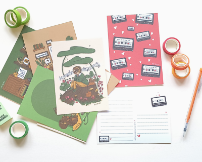 Cartões postais ilustrados com temas de natureza e cassetes, fitas adesivas coloridas e caneta sobre mesa branca.