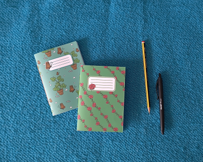 Dois cadernos decorados com padrão de plantas e flores, lápis amarelo e caneta preta sobre tecido azul