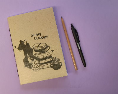 Caderno com capa de papel reciclado e ilustração de gato e livros, lápis e caneta numa superfície lilás.