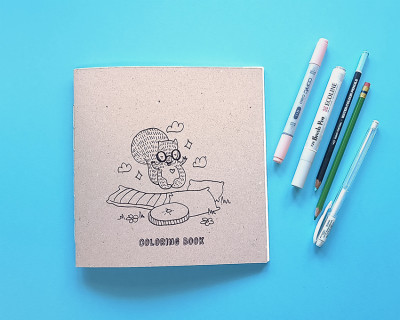 Livro de colorir com desenho de personagem e texto 'COLOURING BOOK' com canetas, lápis e caneta esferográfica ao lado