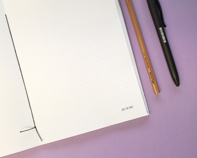 Caderno aberto com páginas pontilhadas, lápis e caneta sobre fundo lilás.