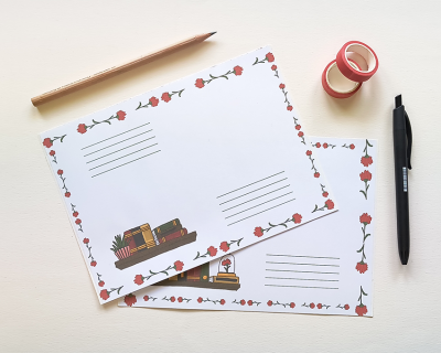 Dois postais decorados com flores e ilustrações de livros sobre mesa branca com lápis, caneta e fita adesiva