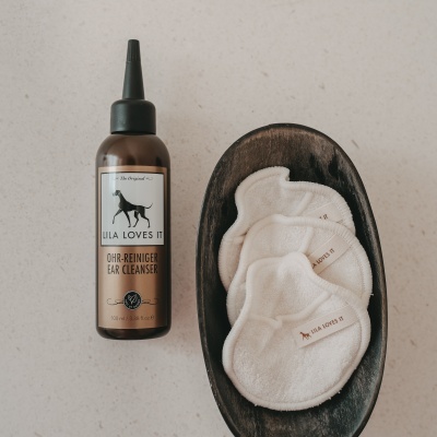 Produto de limpeza auricular para cães e discos de tecido branco numa tigela de madeira.