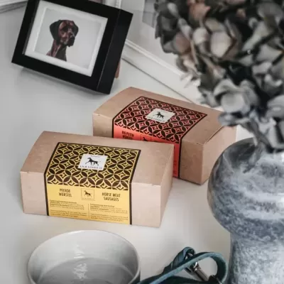 Pacotes de salsichas de carne de cavalo em caixas de papelão com etiquetas decorativas amarelas e vermelhas sobre mesa com frasco, quadro e prato.