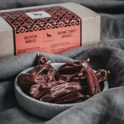 Pequenas salsichas escuras numa tigela cinzenta e embalagem de cartolina cor de rosa e castanha com texto em inglês e alemão.