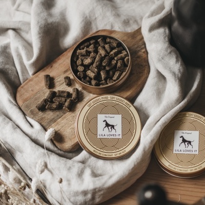 Duas latas de comida para cães LILA LOVES IT em cima de tábua de madeira com comida para cães e tecido bege.