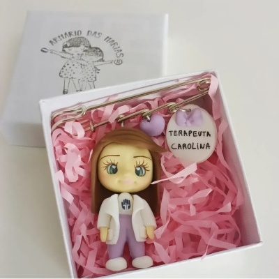 Broche decorativo com figura de boneca e chaveiro com texto, em caixa branca com enchimento rosa