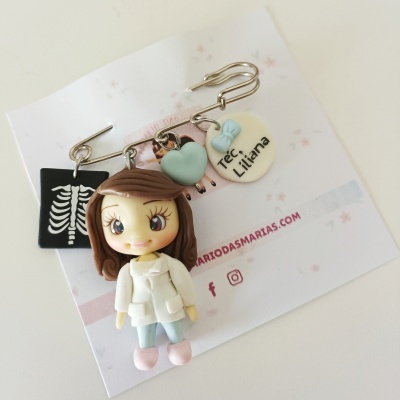 Alfinete de segurança com figuras penduradas incluindo rapariga com bata e texto 'Tec. Liliana'