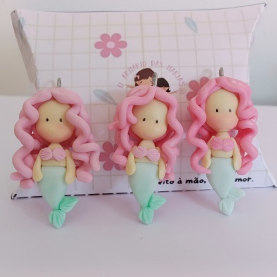 Três figuras pequenas de sereias com cabelo rosa e caudas verdes à frente de uma caixa decorada