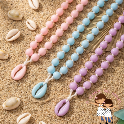 Colares com contas grandes cor-de-rosa, azul e lilás em areia com conchas naturais e artificiais.